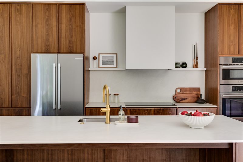 Walnut kitchen seen across the island, with a brass faucet, plaster hood and stainless refrigerator